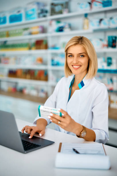 薬局で働きながらカメラを見ながらノートパソコンを使う幸せな女性薬剤師。 - prescription medicine pharmacy medical vertical ストックフォトと画像
