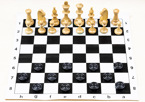 playing by different rules on the same board - black checkers and white chess figures on black white chessboard, chess pieces and checker discs in starting positions (focus on front checkers)