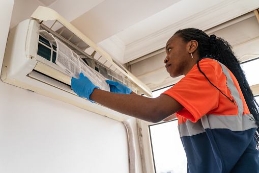 black Female technician service open air conditioner indoor for checking and repairing, Air conditioner service indoors. Air conditioner cleaning technician she opened the front cover and took out the filters and washed it