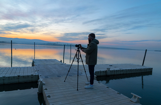 A matured man photographer is looking through the viewfinder and takes photographs of lake at sunrise time, with a tele photo lens mounted on tripod.