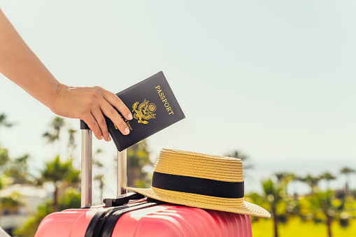 Woman with pink suitcase and amerian passport standing on passengers ladder of airplane opposite sea coastline with palm trees. Tourism concept