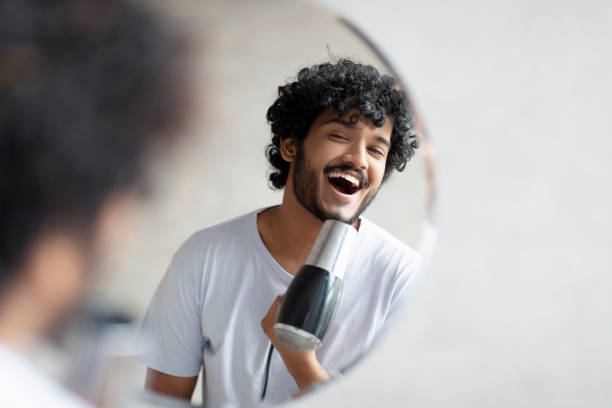 istock Joyful indian man holding hairdryer and singing as in microphone, having fun while doing morning beauty routine 1464830271