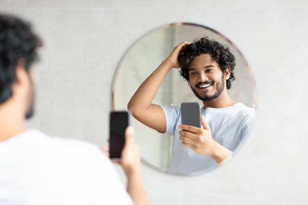 istock Cheerful indian man standing next to mirror in modern bathroom, holding smartphone while doing morning routine 1464830281