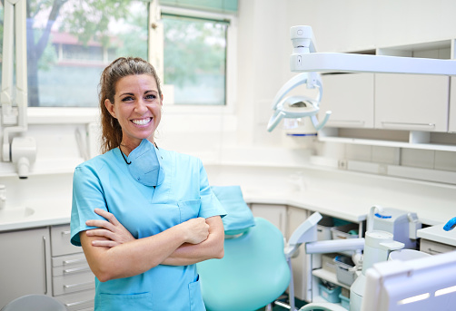 Optimistic woman in blue uniform with mask crossing arms and looking at camera with smile while working in modern office of dental clinic