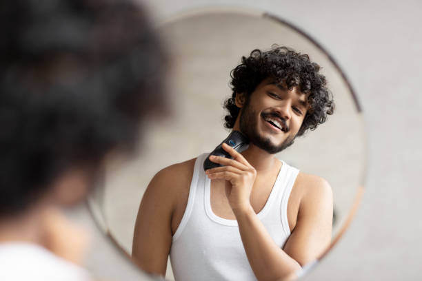 istock Grooming concept. Handsome young indian man looking at mirror and shaving beard with trimmer or electric shaver 1464831068