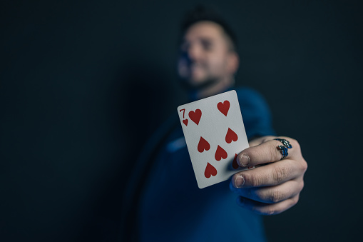 Faded image of a young magician in the dark showing a card with his hand while looking at the camera. The displayed card is in focus.