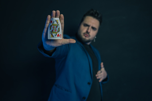 Blurred portrait of a young magician in the dark showing in the palm of his hand a hidden card while looking astonished after the trick. The displayed card is in focus.