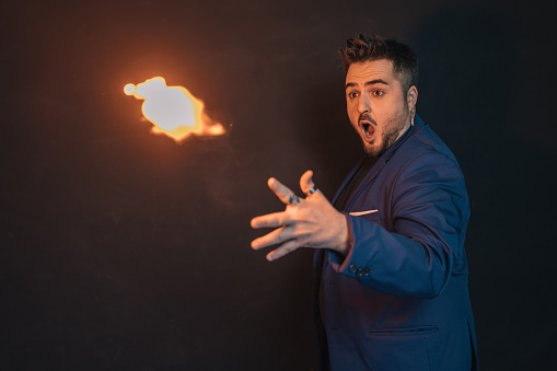 Young magician in the dark with a surprised and dumbfounded face throwing a flaming fireball with one hand.