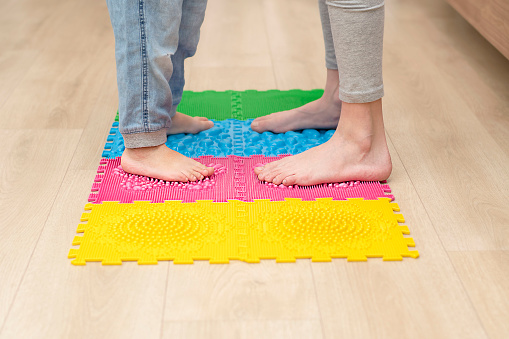 Health concept. Two boys stand barefoot on a colorful orthopedic massage mat in a home interior. Massage and improve circulation of the feet. Close-up of children's legs.