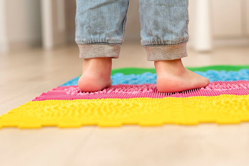Health concept. A little boy stands with bare feet on a multi-colored orthopedic massage mat in a home interior. Close-up of children's heels. Back view.
