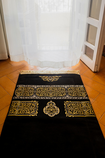 Islamic black prayer rug  with a golden decoration, rug on the floor in front of a window with white curtains, spiritual mood, Ramadan vibes, Ramadan background with copy space