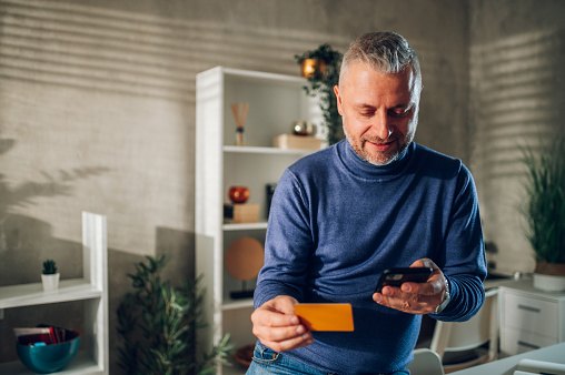 Portrait of middle aged man using smartphone and a credit card while sitting on a table at home. Handsome middle aged guy is online shopping while relaxing at home. Online banking and paying bills.