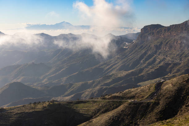 Landsape photo of winding road in tho Gran Canaria's moutains with Tenerifa's Teide view. Daylight, some clouds, and some traffic. No people.