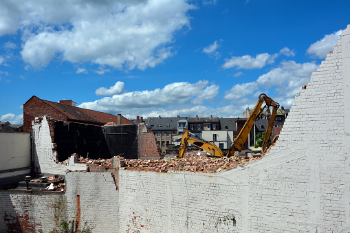 Behind the dilapidated brick wall of the building, excavators are demolishing the house against the background of a bright sky