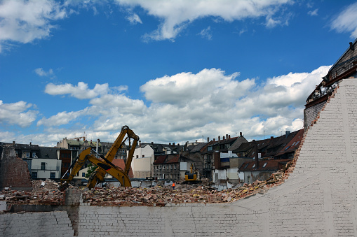 In an ancient city, on the street, excavators dismantle a house behind a wall against the background of a bright sky