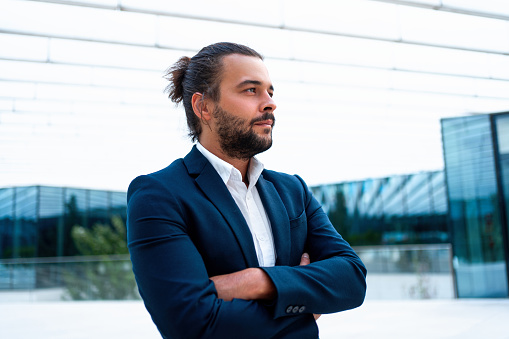 Successful businessman in suit with beard standing in front of office glass building arm crossed confidently looking away. Hispanic male business person side view portrait. Free space. Hipster man