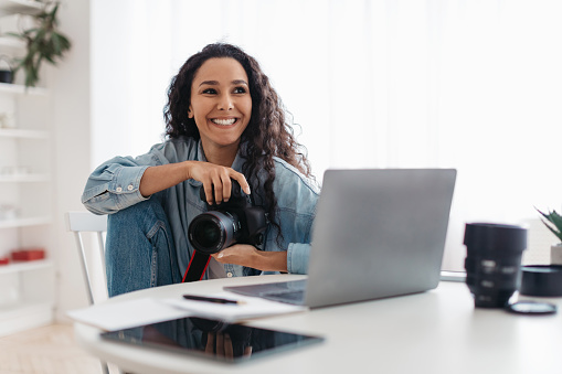 Feliz fotógrafa mujer sosteniendo la cámara fotográfica usando la computadora portátil sentado en el interior photo