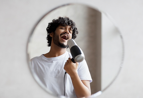 Joyful indian man having fun in the morning in bathroom, holding hairdryer and singing as in microphone, doing morning beauty routine, copy space