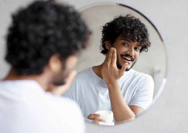 istock Positive indian man applying moisturizer cream on cheeks, holding cosmetic jar while standing near mirror in bathroom 1464866892