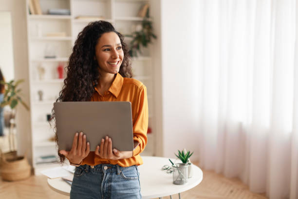 woman holding laptop working online standing looking aside smiling indoor - aside imagens e fotografias de stock
