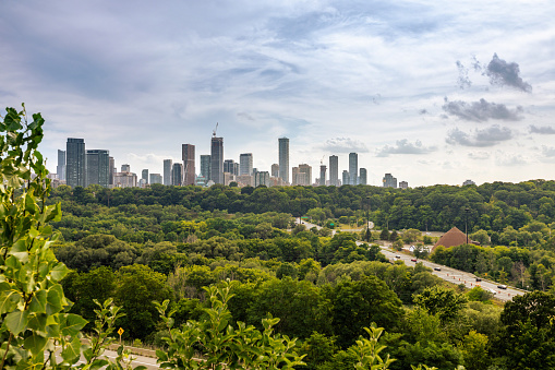 This 7 August 2022 daylight photo shows the downtown skyline of Toronto, Ontario, Canada. The foreground of lush foliage along the Don River and nearby parkland transition sharply to skyscrapers in the distance. Traffic to and from Don Valley Parkway is seen on the right. Numerous construction cranes are seen along the cityscape.