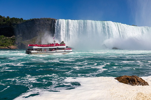 This 12 August 2022 daytime photo shows the Niagara Wonder (O.N. 837992), a tourist boat, making its way upstream along the turbulent Niagara River toward the centre of the Horseshoe Falls in Niagara Falls, Ontario, Canada. Sightseers dressed in red ponchos line the decks of the boat. In the background are the Horseshoe Falls with tourists at the top on the United States side.