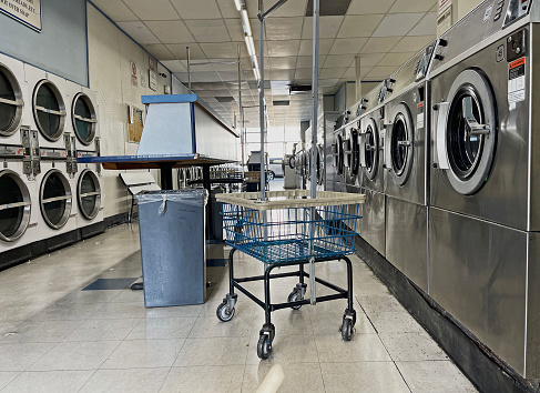 Interior of a laundromat.   Clothes dryers along left wall and washers on right side of photograph.   A blue, clothes basket with wheels, tables, and trash can.