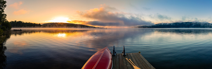 This August 2022 panoramic image shows the sunrise at Lake of Two Rivers in Algonquin Provincial Park, Ontario, Canada. A red canoe with wooden oars sits on a dock in the foreground. In the background, the morning sun rises over a forested hill. Mist hugs the surface of the lake, as low clouds reflect multiple colors in the dawn sky.