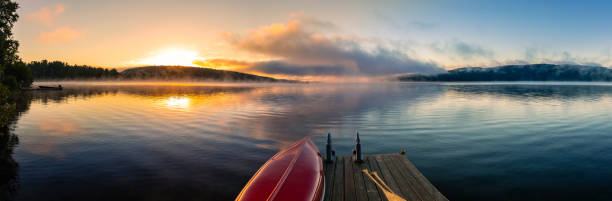 Lake of Two Rivers, Algonquin Provincial Park, Summer Sunrise This August 2022 panoramic image shows the sunrise at Lake of Two Rivers in Algonquin Provincial Park, Ontario, Canada. A red canoe with wooden oars sits on a dock in the foreground. In the background, the morning sun rises over a forested hill. Mist hugs the surface of the lake, as low clouds reflect multiple colors in the dawn sky. provincial park stock pictures, royalty-free photos & images