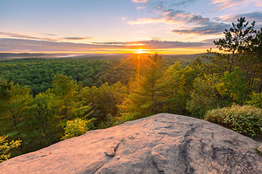 This August 2022 image shows the sun setting over Algonquin Provincial Park, Ontario, Canada. A rocky cliff at the top of the Lookout Trail provides an impressive setting to take in the forested landscape.