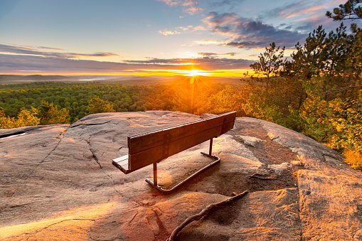 This August 2022 image shows the sun setting over Algonquin Provincial Park, Ontario, Canada. A bench on a rocky cliff at the top of the Lookout Trail provides an impressive setting to take in the forested landscape.