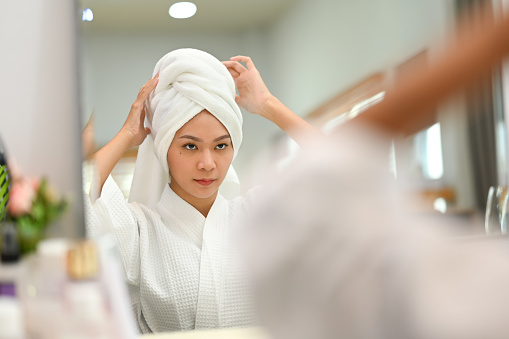 Smiling millennial woman in bathrobe standing front of mirror and making daily beauty routine at home.