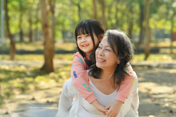 Smiling middle age grandmother giving piggyback ride to cute little granddaughter during walking on in the park. stock photo