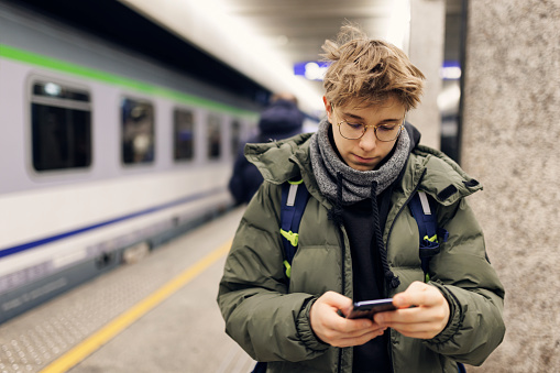 Teenage boy standing in the train station on the railroad platform. The boy is checking the train time in the smartphone.\nCanon R5