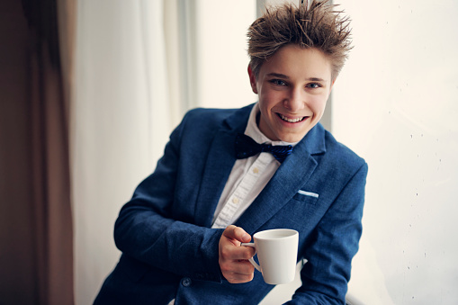Portrait of a cute teenage boy wearing a suit and a bow tie. The boy is looking at the city and holding a cup of coffee.\nCanon R5