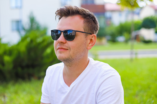 Young male man portrait. European caucasian white young student in sunglasses on green grass lawn on town street, park. Lifestyle Headshot portrait, tourist, businessman