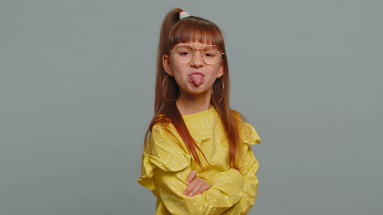 Cheerful funny bully young toddler girl in glasses showing tongue making faces at camera, fooling around, joking, aping with silly face, teasing. Preteen child kid on gray background. Little children