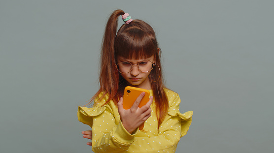 Young toddler school girl in glasses use smartphone typing browsing, loses becoming surprised sudden lottery results, bad fortune, loss. Education, study. Teen female child kid on gray background