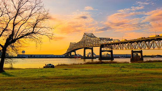 Horace Wilkinson Bridge over the Mississippi River green riverbank park with a barren willow tree at sunrise with golden glowing clouds on the blue sky in Baton Rouge, Louisiana, USA