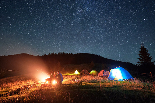 Night camping in mountains under starry sky. Silhouette of two people, couple sitting on grass near campsite, admiring landscape. Concept of tourism and healthy living.