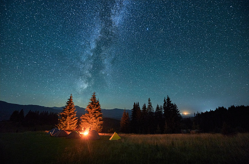 Night camping in mountains under starry sky. Tourist tents in campsite under sky full of stars with Milky way. Silhouette of hikers having a rest near burning campfire. Concept of tourism, adventures.