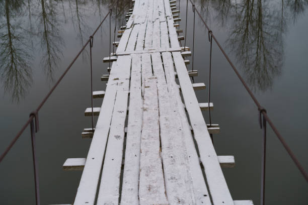 Photography of snowy suspended wooden bridge above river Desna in russian countryside. Gloomy day. White and gray colors. Touristic and lifestyle concepts. Fish eye lens.