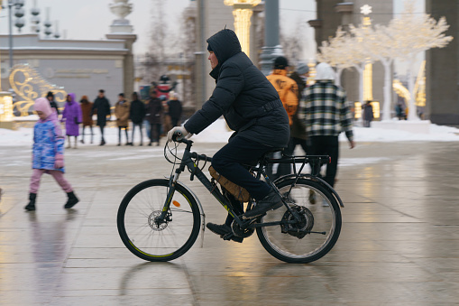 Moscow, Russia - December 15, 2022: Young man drives bicycle on the city street in winter day. VDNkH public park. Road is snowy and wet. He is cold. Side view. Fast transportation theme