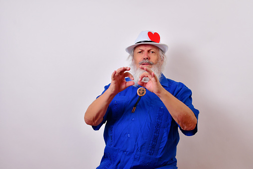An old man celebrating Valentine's day with a red paper heart on his hatband looking serious and making pay attention sign with his hands