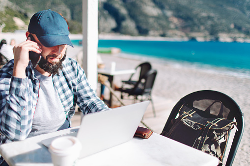 Young adult man working remote from beach cafe and smoking