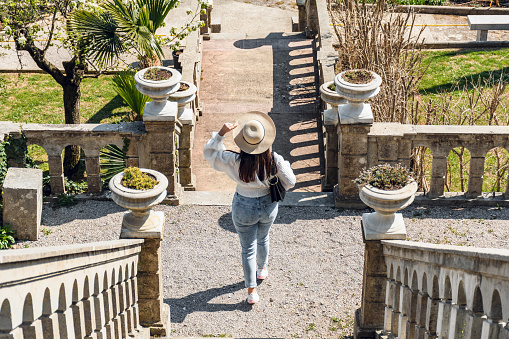 Back view of young woman in jeans and white blouse standing in beautiful mediterranean gardens on sunny day in spring, summer.