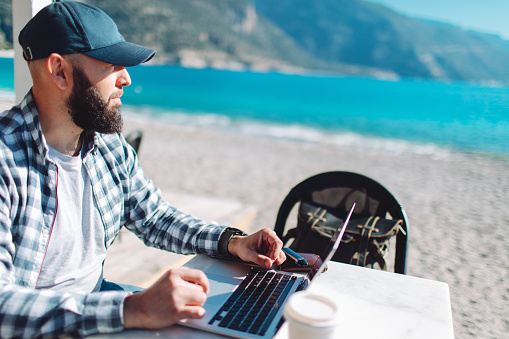 Young adult man working from beach cafe using laptop