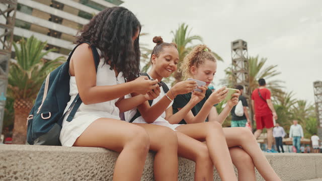 Three girls friends pre-teenage are sitting on the waterfront using mobile phone. Three teenagers playing games on a smartphone on the outdoors in urban cityscape background