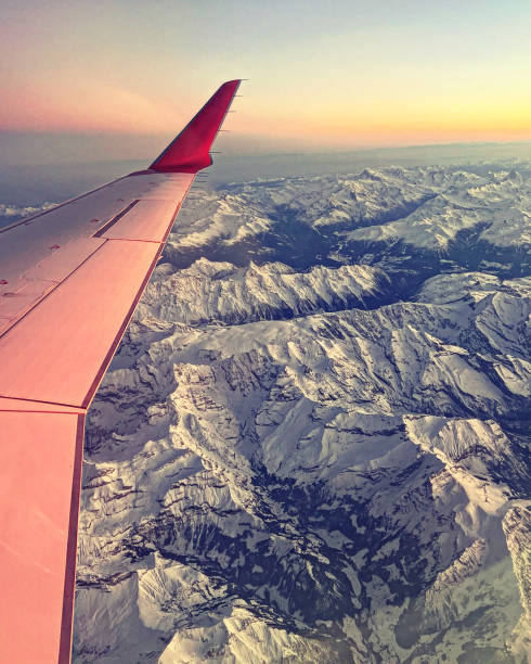 Bird’s eye view (from a window seat) of the Alps during sunset with beautiful pink colours of the sun. The wing of the airplane is visible on the left hand side of the frame.