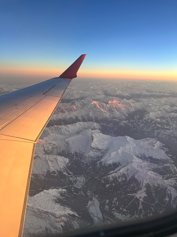 Bird’s eye view (from a window seat) of the Alps during sunset with beautiful pink colours of the sun. The wing of the airplane is visible on the left hand side of the frame.